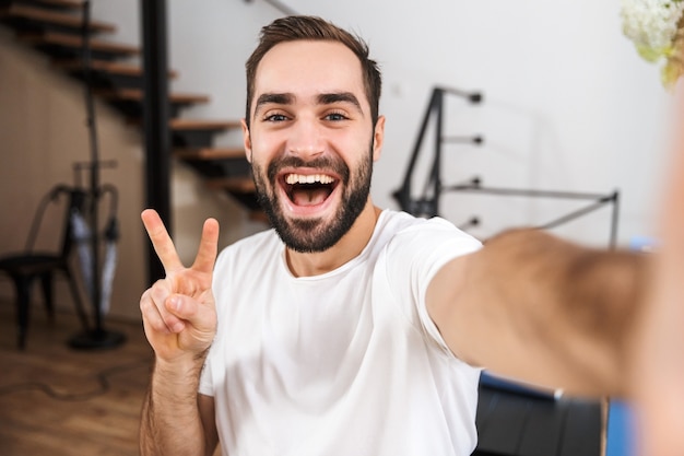 Uomo felice che prende un selfie mentre era seduto in cucina a casa, gesto di pace