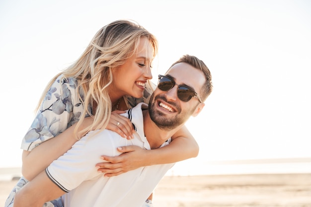 uomo felice con la barba lunga che indossa occhiali da sole sorride e fa un giro sulle spalle donna seducente mentre cammina sulla spiaggia soleggiata