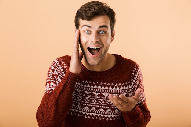 uomo felice con stoppie che indossa un maglione lavorato a maglia urlando e toccando il viso, isolato sul muro beige