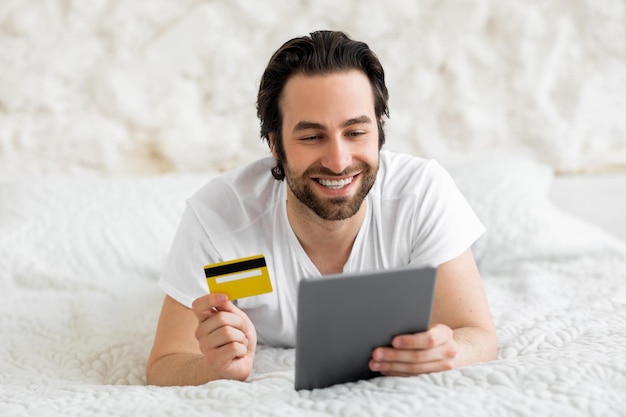 Uomo felice con tavoletta digitale e carta di credito a letto
