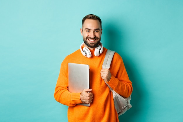 Uomo felice con zaino e cuffie, in possesso di laptop e sorridente, andando a lavorare, in piedi sopra