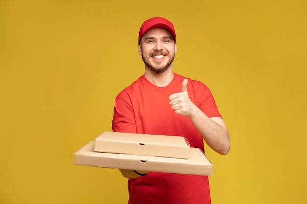 Uomo felice dal servizio di consegna in maglietta rossa e cappuccio che danno l'ordine del cibo e che tiene la scatola della pizza isolata sopra la parete gialla