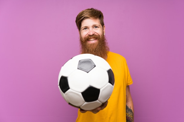 Uomo felice di Redhead con la barba lunga che tiene un pallone da calcio sopra la parete porpora