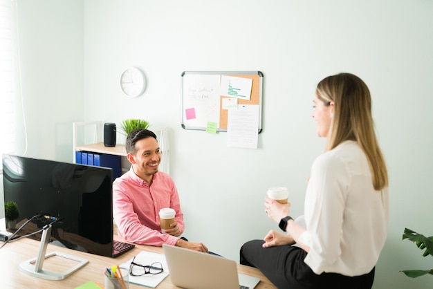 Uomo felice e donna caucasica sorridente seduti alla loro scrivania mentre parlano di pettegolezzi in ufficio. Colleghi professionisti che prendono una pausa dal lavoro e bevono caffè