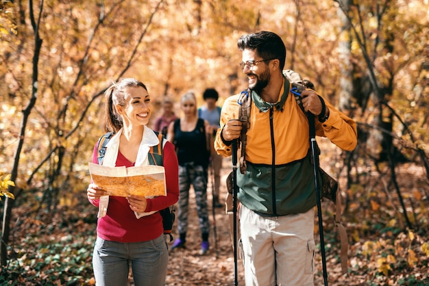 Uomo felice e donna che fanno un'escursione nei boschi in autunno. Mappa della tenuta della donna ed uomo che la esaminano e che sorridono. Sullo sfondo altri escursionisti a piedi.