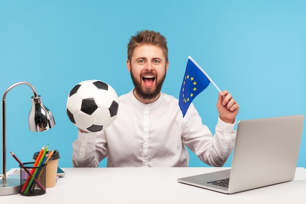 Uomo felice e soddisfatto che tiene in mano un pallone da calcio bianco e nero e la bandiera dell'UE che va a guardare il calcio UEFA Champions League e supporta la squadra preferita Colpo in studio indoor isolato su sfondo blu