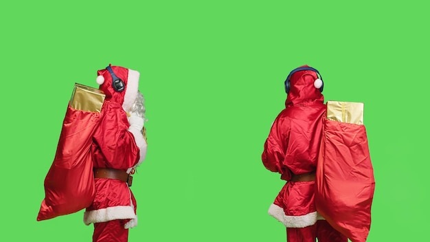 Uomo felice in costume da Babbo Natale che balla, ascolta musica con un sacco di regali su schermo verde. Allegro personaggio invernale che si diverte con l'auricolare vestito con il famoso abito rosso con barba bianca.