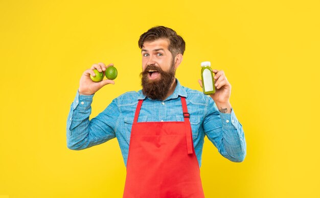 Uomo felice in grembiule con lime e bottiglia di succo su sfondo giallo, barista di succhi.