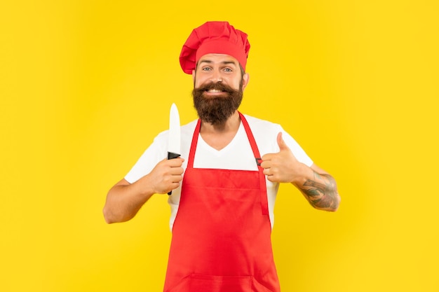 Uomo felice in grembiule rosso e toque dando pollice in possesso di chef coltello sfondo giallo, chef