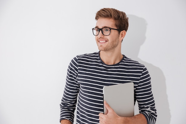 Uomo felice in maglione a righe che tiene in mano il portatile e distoglie lo sguardo. Sfondo grigio isolato