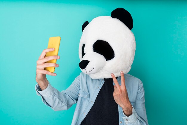 Uomo felice in maschera panda che si fa un selfie con lo smartphone mentre gesticola il segno della vittoria sul muro blu