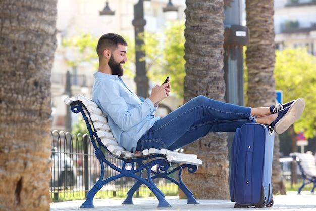 Uomo felice integrale con cellulare e valigia seduto sulla panchina