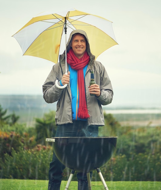 Uomo felice ombrello e barbecue sotto la pioggia con birra per la cena o la cena sulla griglia del fuoco Persona di sesso maschile che sorride per la copertura assicurativa o la protezione durante la cottura sotto la pioggia o il maltempo