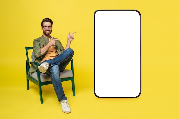 Uomo felice seduto in poltrona e che punta al cellulare gigante con mockup di schermo bianco vuoto per annunci online