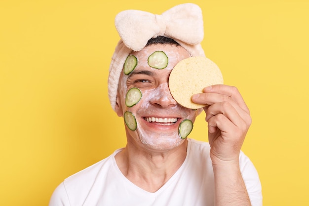 Uomo felice sorridente che indossa maglietta bianca e fascia per capelli con fiocco in posa con maschera facciale e cetriolo che esprime felicità che copre l'occhio con spugna isolata su sfondo giallo