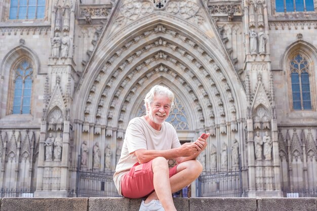 Uomo felice, viaggiatore anziano dai capelli bianchi che si gode la visita alla cattedrale di Barcellona, seduto su una scala che tiene il telefono cellulare