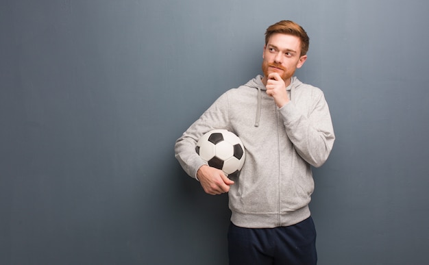 Uomo fitness giovane rossa dubbioso e confuso. Sta tenendo un pallone da calcio.