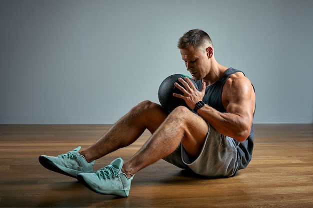 Uomo forte che fa esercizio con la palla medica. Foto del fisico perfetto dell'uomo sulla parete grigia. Forza e motivazione.