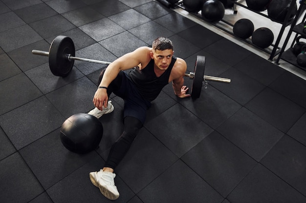 Uomo forte in abiti sportivi seduto sul pavimento in palestra con attrezzature