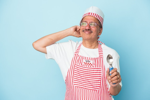 Uomo gelato indiano senior che tiene una paletta isolata su fondo blu che tocca la parte posteriore della testa, pensando e facendo una scelta.