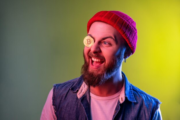 Uomo giocoso hipster in piedi che copre un occhio con la criptovaluta bitcoin dorata che guarda da parte