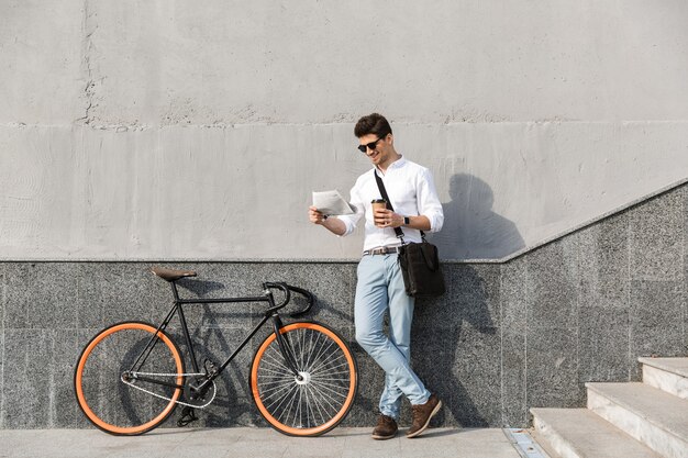 uomo gioioso che indossa occhiali da sole, beve caffè da asporto e legge il giornale, stando in piedi con la bicicletta lungo il muro all'aperto