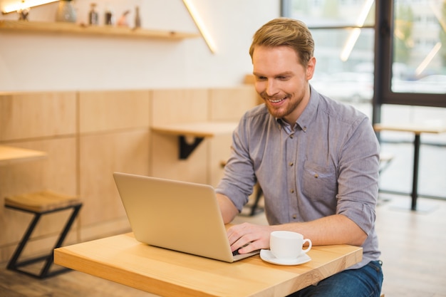 Uomo gioioso felice positivo che si siede nella caffetteria e lavora al computer portatile mentre si gode il suo lavoro