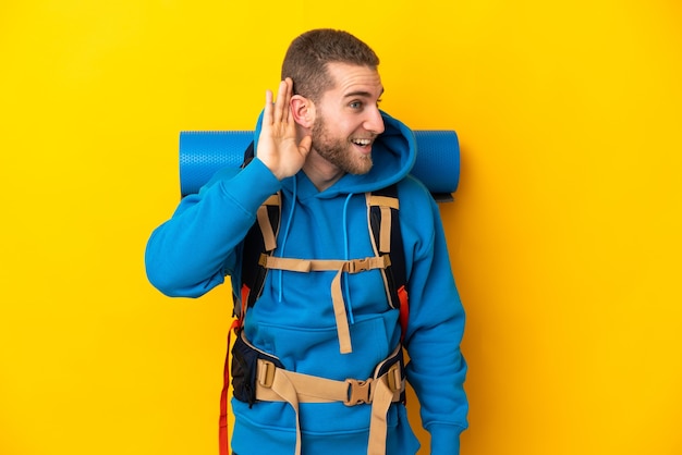 Uomo giovane alpinista caucasico con un grande zaino isolato su giallo ascoltando qualcosa mettendo la mano sull'orecchio