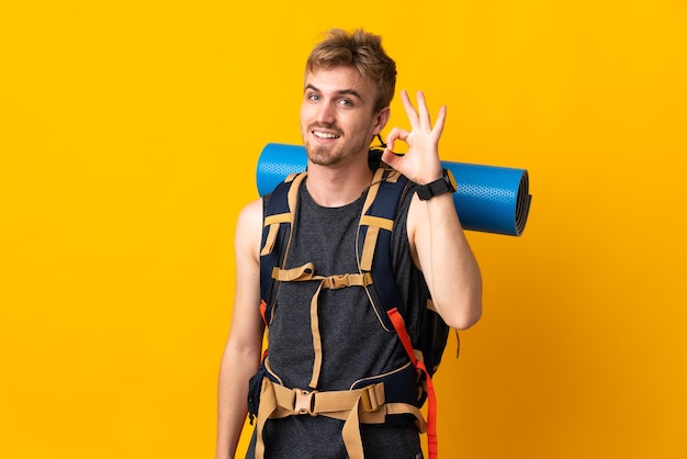 Uomo giovane alpinista con un grande zaino isolato su sfondo giallo che mostra segno ok con le dita