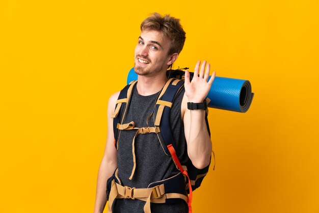 Uomo giovane alpinista con un grande zaino isolato su sfondo giallo salutando con la mano con l'espressione felice