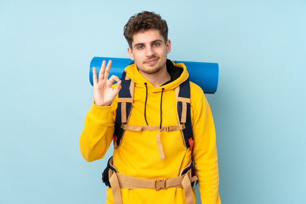 Uomo giovane alpinista con un grande zaino sul blu che mostra un segno giusto con le dita