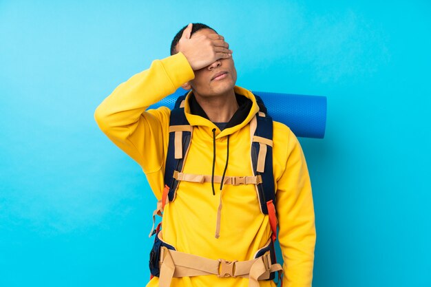 Uomo giovane alpinista con un grande zaino sul muro blu isolato che copre gli occhi a mano