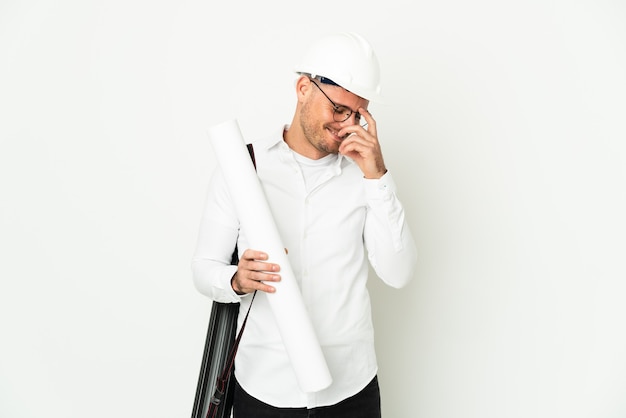 Uomo giovane architetto con casco e azienda schemi isolati su sfondo bianco ridendo