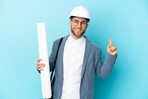 Uomo giovane architetto con casco e in possesso di schemi sulla parete isolata che mostra e alza un dito in segno del meglio