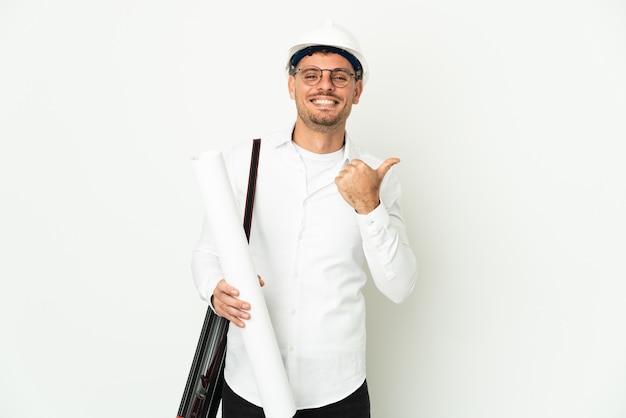 Uomo giovane architetto con casco e tenendo le cianografie isolate sul muro bianco che punta di lato per presentare un prodotto