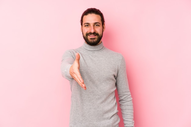 Uomo giovane capelli lunghi isolato su uno sfondo rosa che allunga la mano alla macchina fotografica nel gesto di saluto.