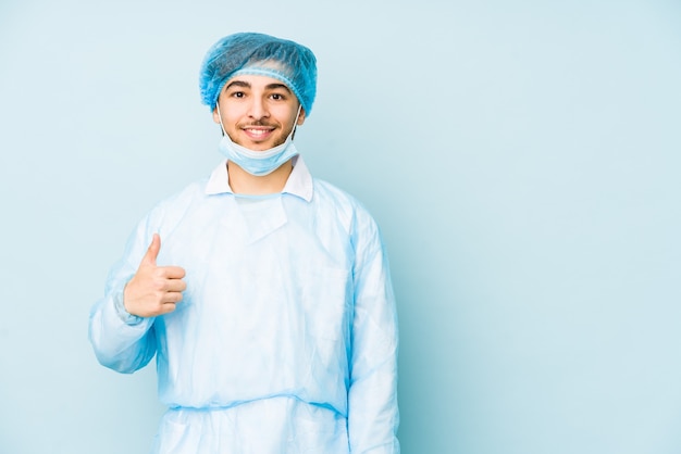 Uomo giovane chirurgo arabo isolato contro su uno sfondo blu sorridendo e alzando il pollice