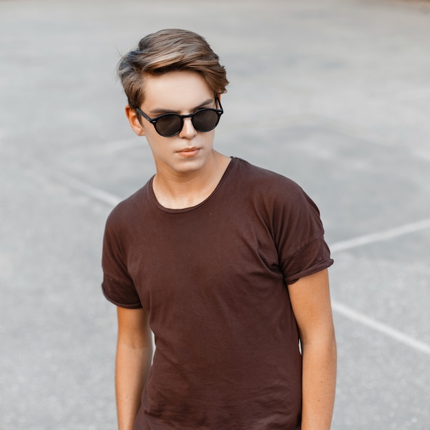 Uomo giovane hipster alla moda bello con un'acconciatura alla moda in una maglietta marrone alla moda in occhiali da sole alla moda sul campo da basket