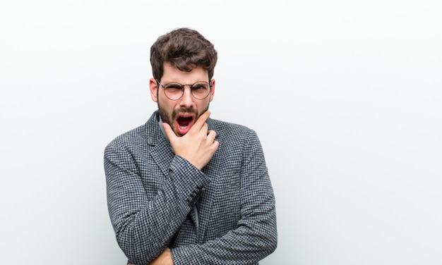 Uomo giovane manager con la bocca e gli occhi spalancati e la mano sul mento, sentendosi spiacevolmente scioccato, dicendo cosa o wow contro il bianco