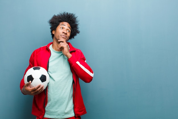 Uomo giovane sportivo nero con un pallone da calcio contro la parete blu del grunge