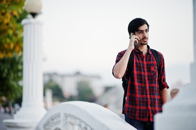 Uomo giovane studente a camicia a scacchi e jeans con zaino parlando sul telefono cellulare