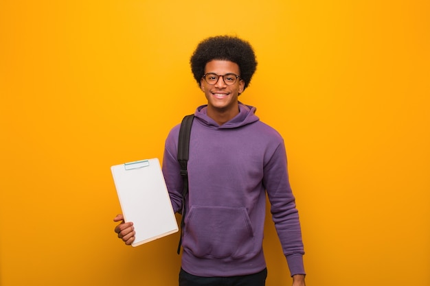 Uomo giovane studente afroamericano che tiene una lavagna per appunti allegra con un grande sorriso