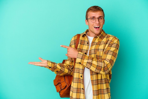 Uomo giovane studente caucasico isolato su blu eccitato in possesso di uno spazio di copia sul palmo.