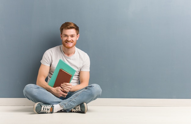 Uomo giovane studente di redhead seduto sul pavimento allegro con un grande sorriso