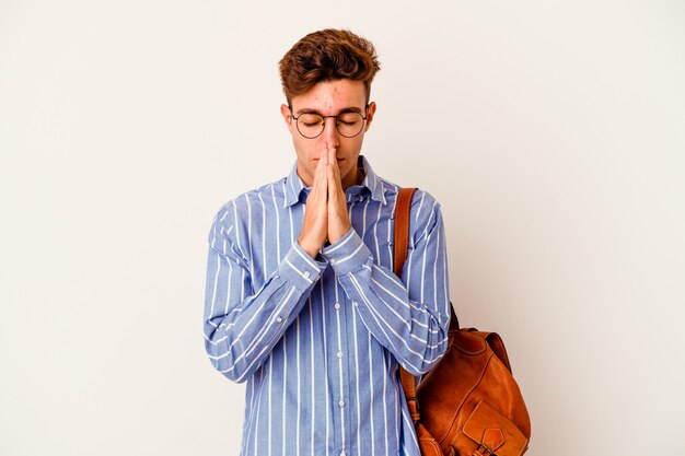 Uomo giovane studente isolato su sfondo bianco tenendo le mani in preghiera vicino alla bocca, si sente fiducioso.