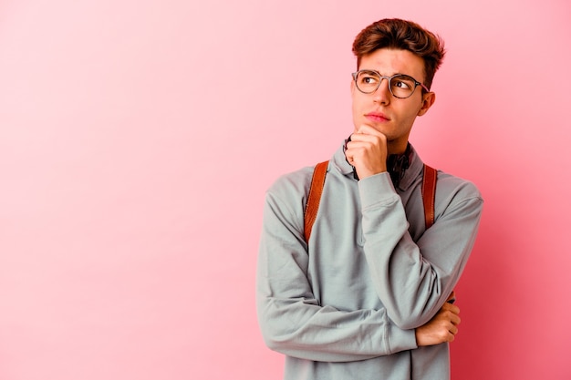 Uomo giovane studente isolato sul muro rosa guardando lateralmente con espressione dubbiosa e scettica