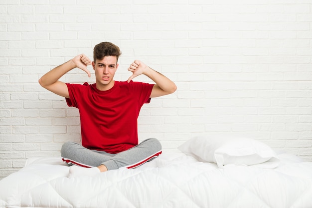 Uomo giovane studente studente sul letto che mostra il pollice verso il basso e che esprime antipatia.