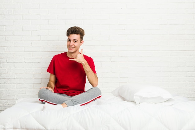 Uomo giovane studente studente sul letto che mostra un gesto di chiamata cellulare con le dita.