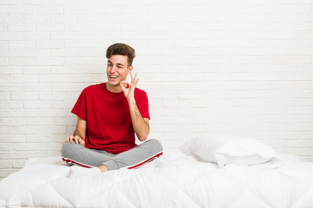 Uomo giovane studente sul letto strizza l'occhio e tiene un gesto ok con la mano.