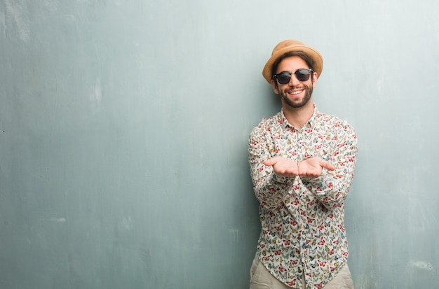 Uomo giovane viaggiatore indossa una camicia colorata in mano qualcosa con le mani, mostrando un prodotto, sorridente e allegro, offrendo un oggetto immaginario